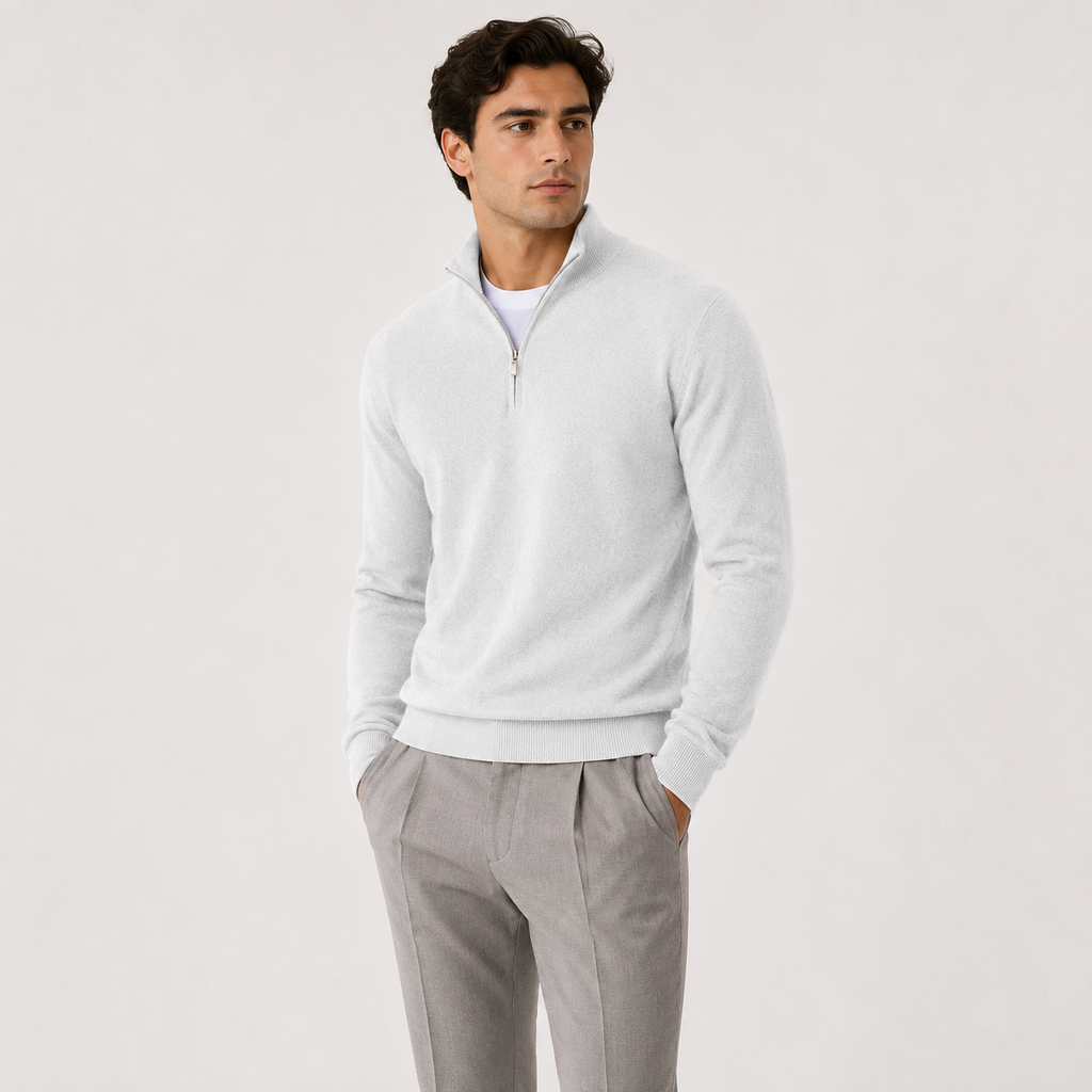 A man wearing a light gray quarter-zip knit sweater over a white undershirt and gray trousers, standing against a plain light gray studio background.