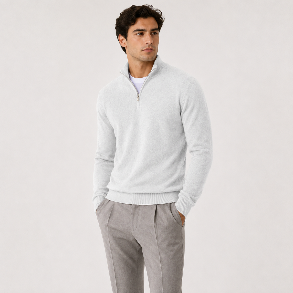 A man wearing a light gray quarter-zip knit sweater over a white undershirt and gray trousers, standing against a plain light gray studio background.