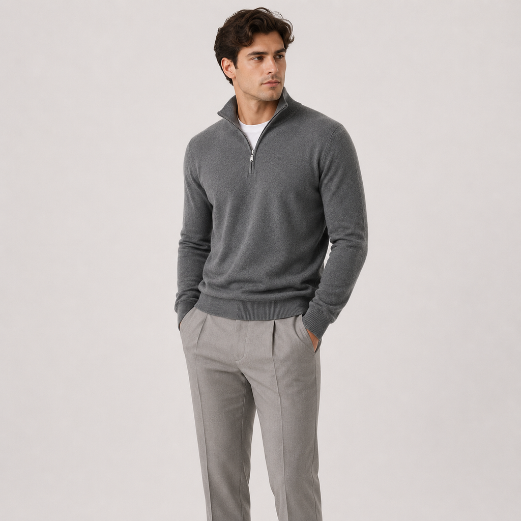 A man wearing a dark gray quarter-zip knit sweater over a white undershirt and light gray trousers, standing against a plain light gray studio background.